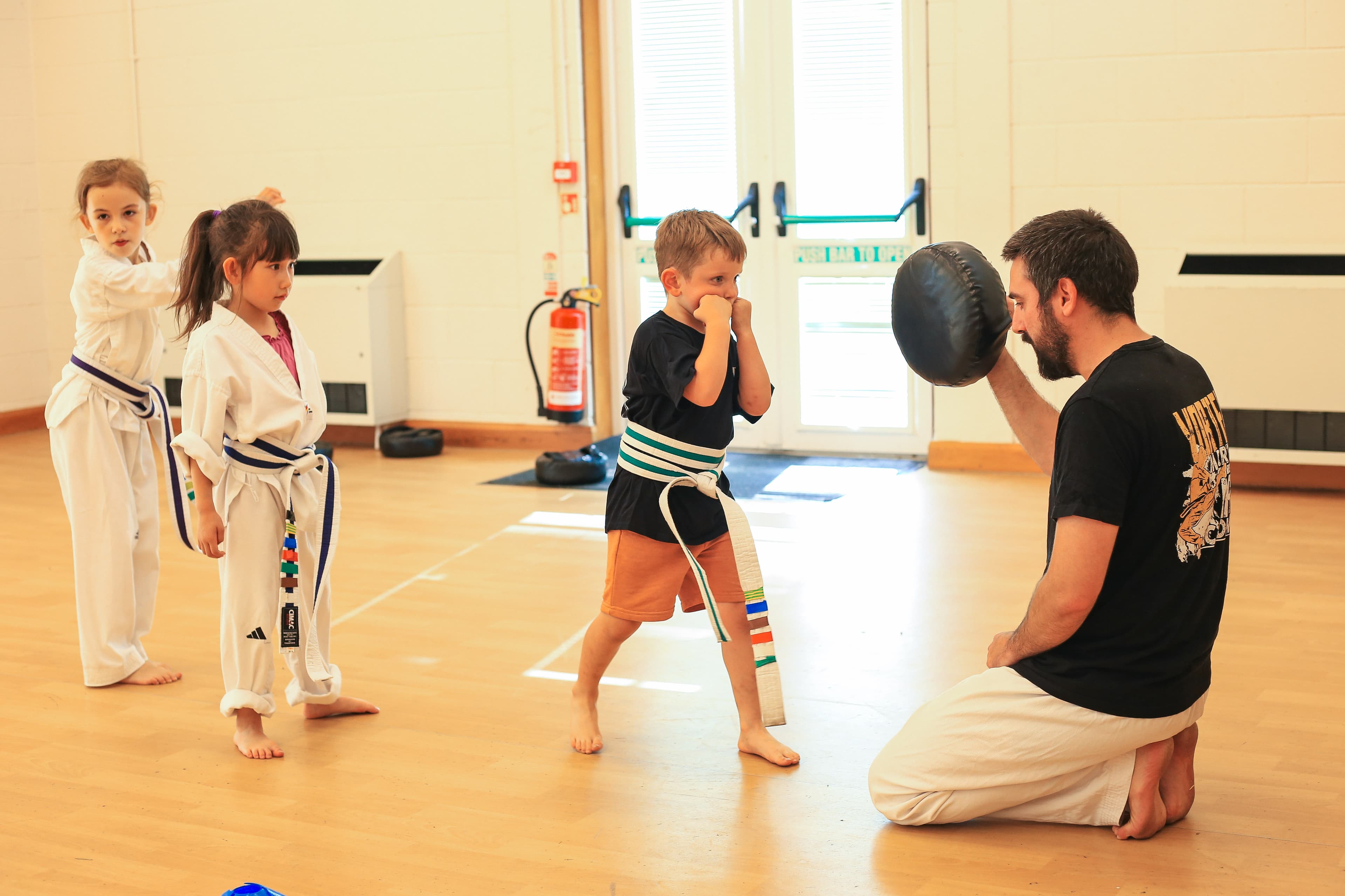 Andy kneeling at a child's level with a pad during a Cadets class at Vortex Martial Arts Academy